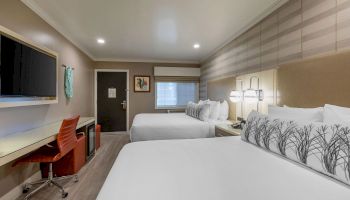 This image shows a hotel room with two beds, a desk with a chair, a flat-screen TV, and modern decor in neutral tones ending the sentence.