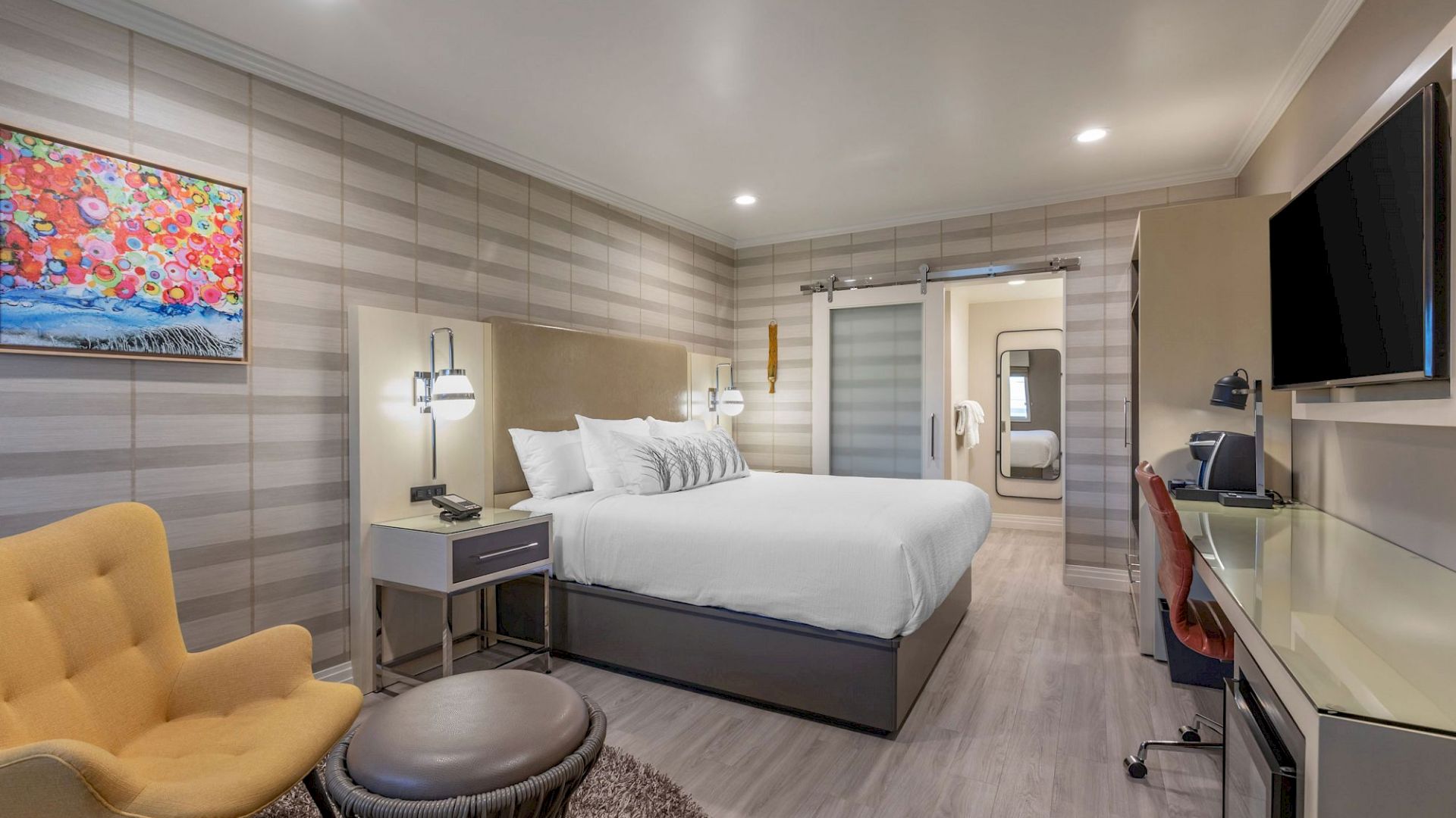 A modern hotel room with a bed, colorful artwork, yellow chair, desk, and television; neutral tones dominate the decor.
