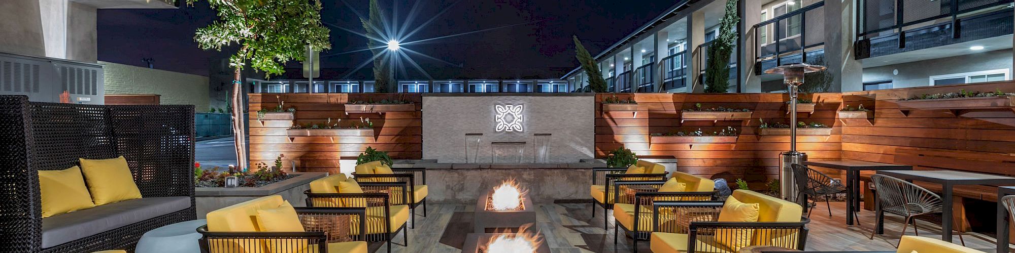 An outdoor patio with yellow cushioned chairs surrounding a rectangular fire pit, next to a modern building, illuminated at night.