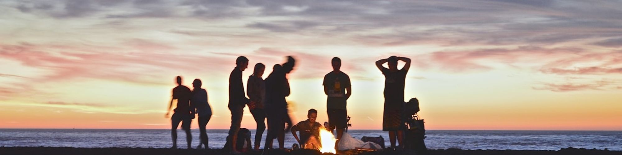 A group of people is gathered around a campfire on a beach during sunset, with the ocean in the background and a colorful sky above.