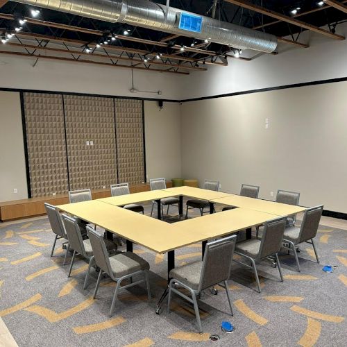 A modern meeting room with a U-shaped table setup, twelve chairs, patterned carpet, exposed ceiling ducts, a small window, and wall decor.