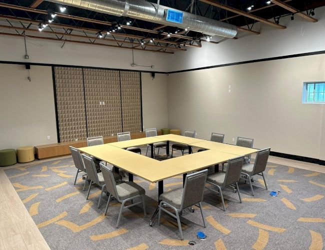 A modern meeting room with a U-shaped table setup, twelve chairs, patterned carpet, exposed ceiling ducts, a small window, and wall decor.