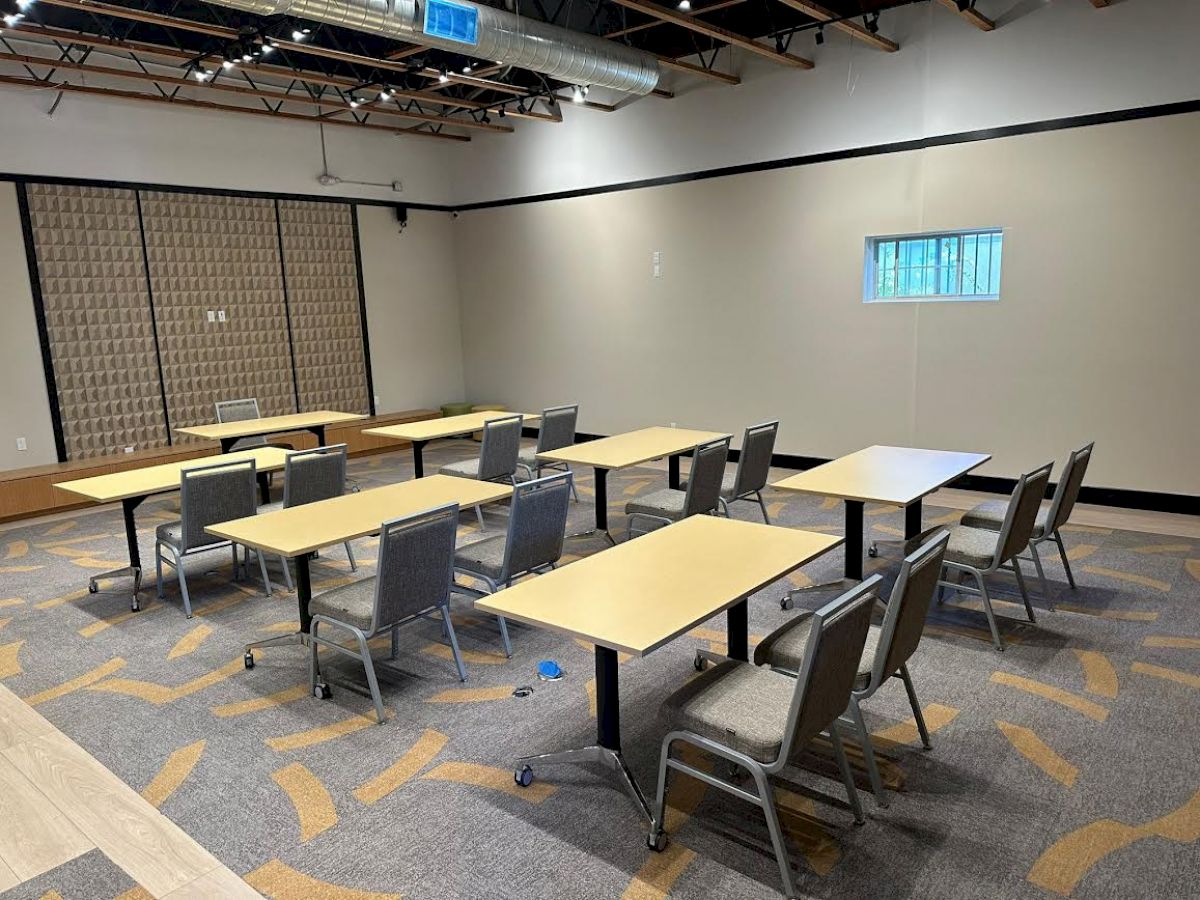 The image shows an empty conference or classroom setup with tables and chairs arranged in a socially distanced manner, within a modern room.