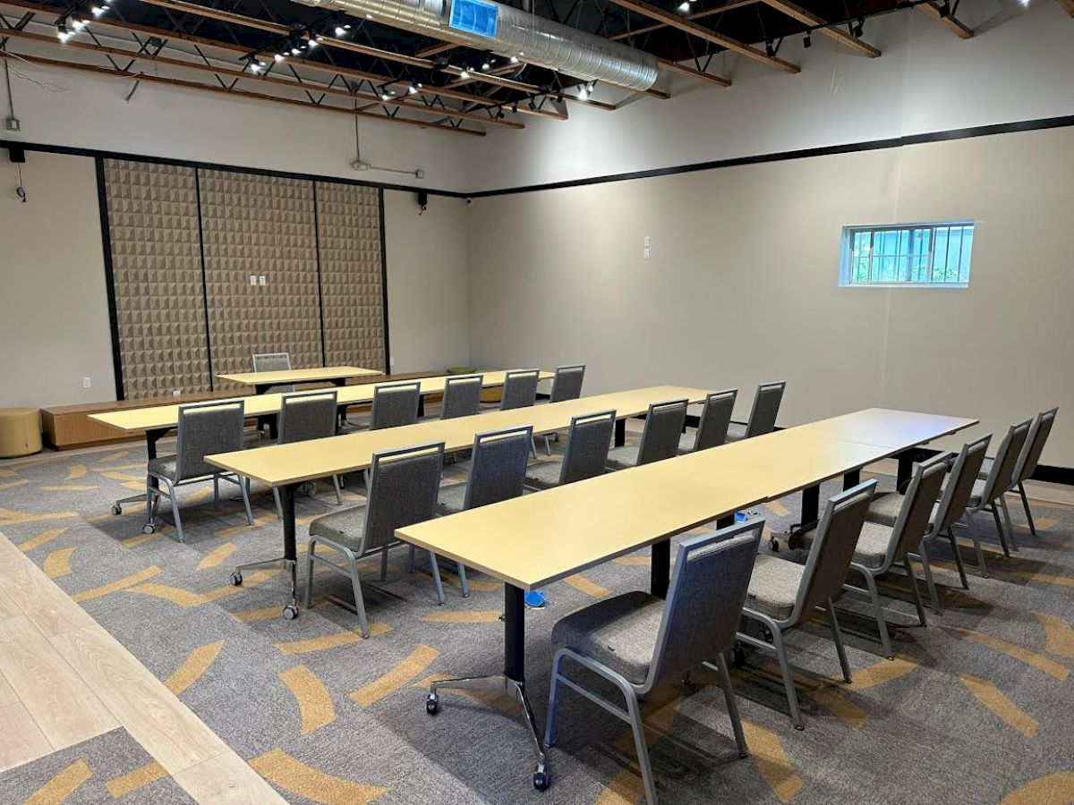 The image shows a conference room with long tables and several chairs arranged in rows, a small window, and a retractable divider on the back wall.