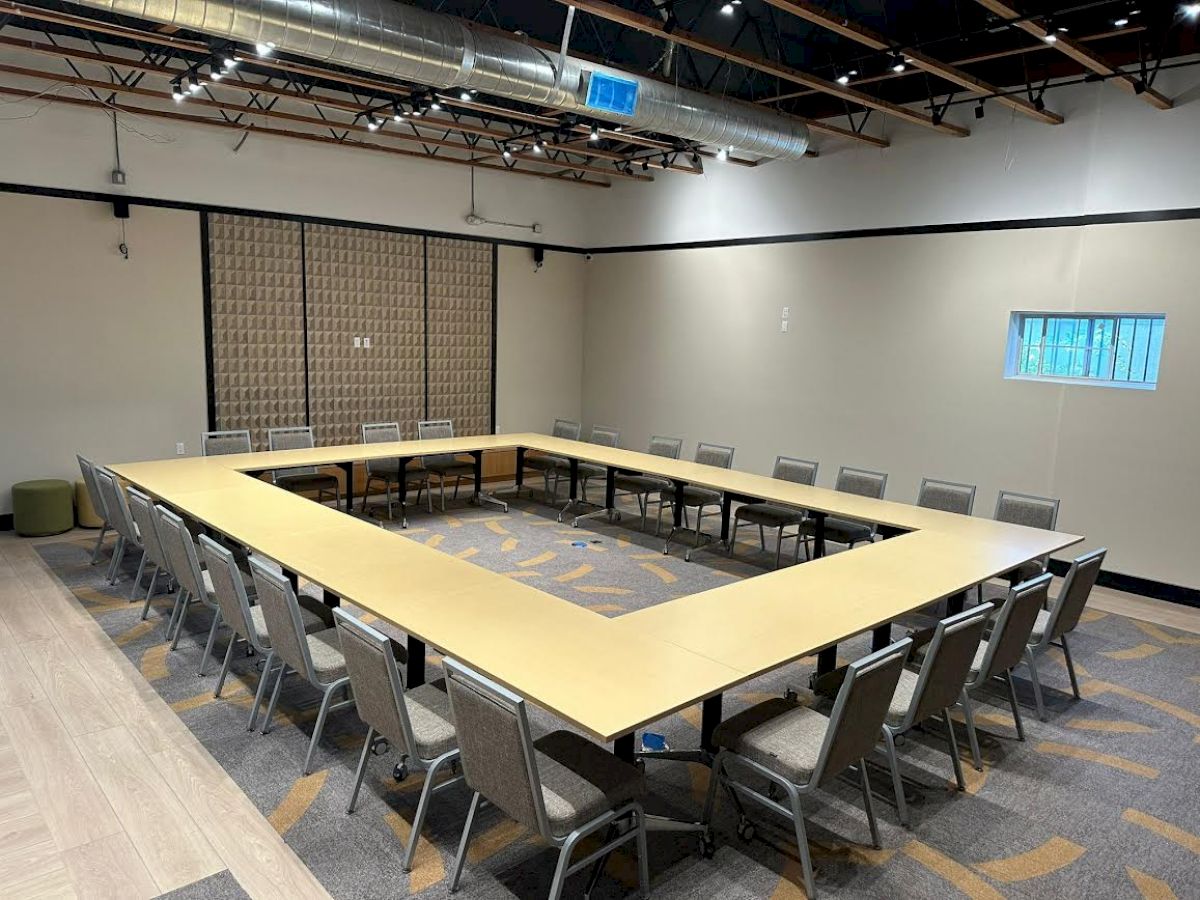 The image shows a conference room with a large, square arrangement of tables and chairs, a carpeted floor, and a small window on one wall.