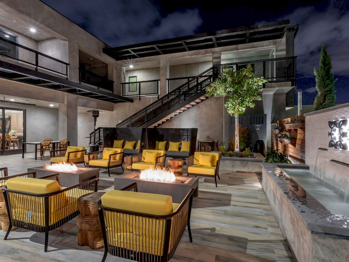 This image shows an outdoor seating area at night, featuring fire pits, yellow chairs, tables, and a modern water feature against an illuminated backdrop.