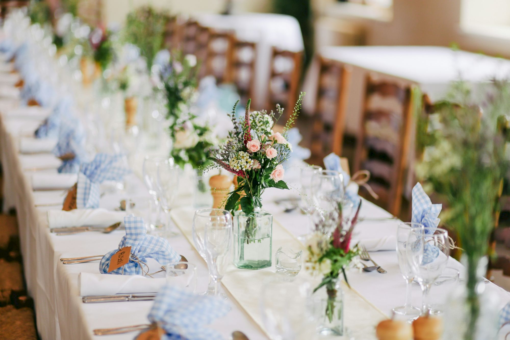 A long, elegantly set table features glassware, floral centerpieces, and checkered napkins, suggesting preparation for a formal meal or event.