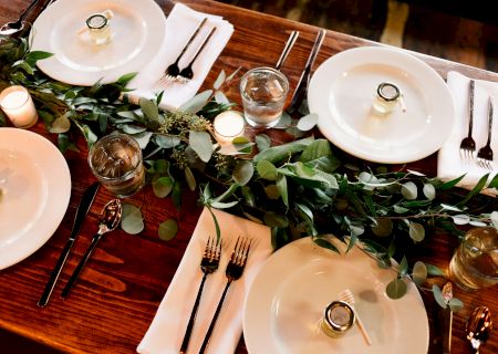 The image shows an elegantly set dining table with plates, cutlery, greenery centerpiece, candles, and small jars on each plate.