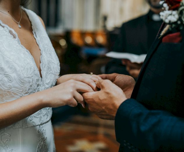 A couple is exchanging rings during a wedding ceremony, with an officiant in the background holding a booklet.