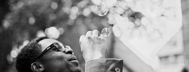 A person wearing glasses and a suit is blowing bubbles outdoors, with a blurred background of trees and people.