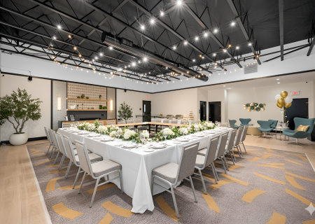 An elegantly decorated event space with a U-shaped table setup, flowers, chairs, modern lighting, and balloons in the background, ready for guests.