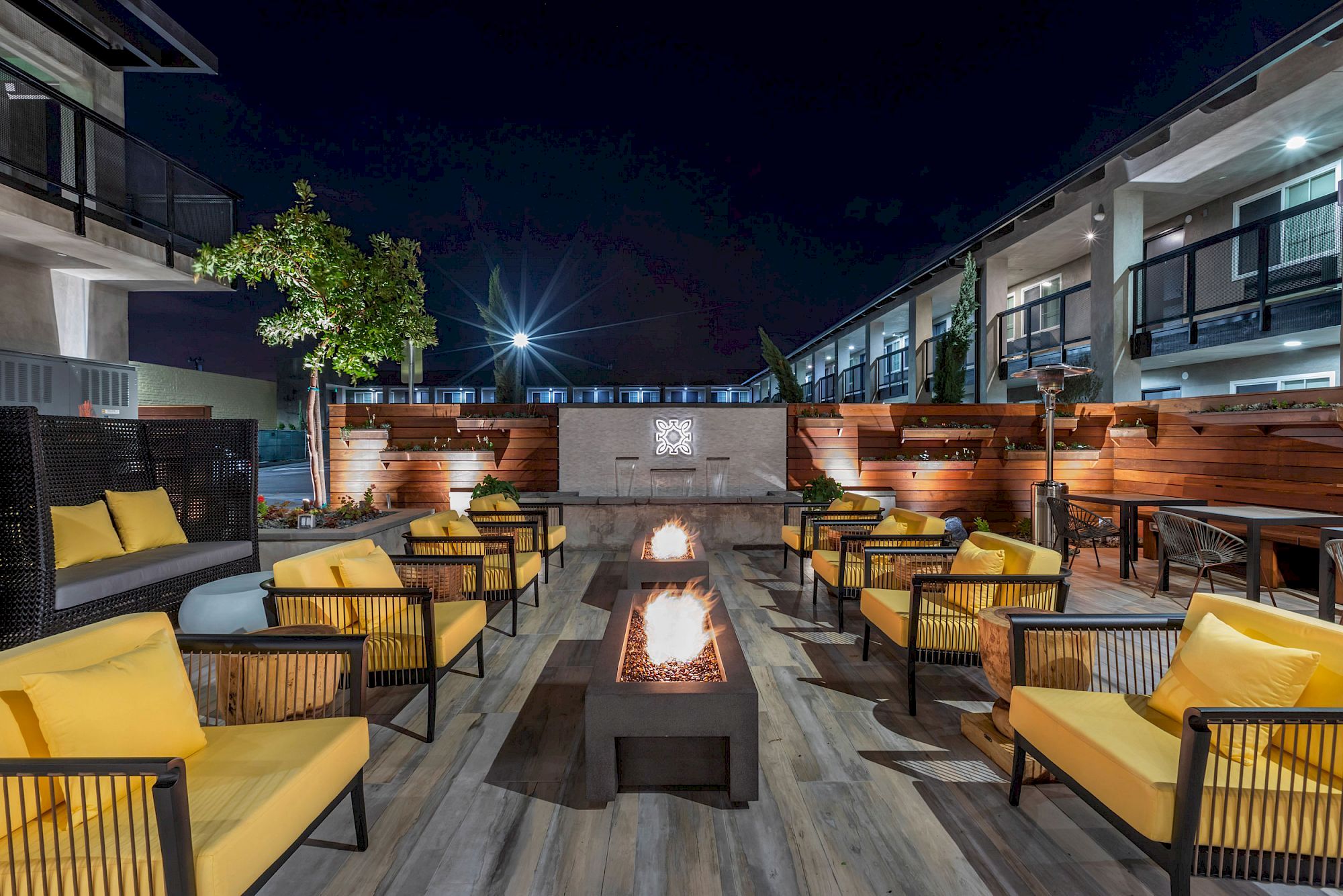 An outdoor lounge area with a central fire pit, yellow cushioned chairs, modern decor, and ambient lighting, set against a backdrop of buildings at night.