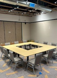 The image shows a modern conference room with tables arranged in a square, surrounded by chairs. The room has a neutral color palette and high ceilings.