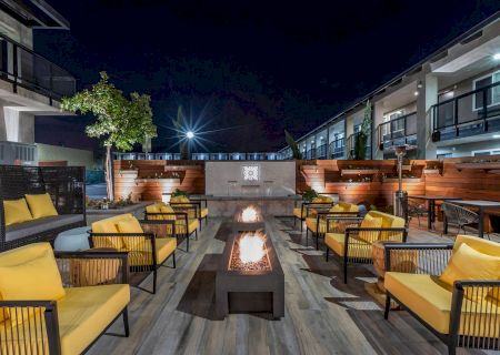 An outdoor lounge area at night with multiple yellow cushioned chairs around two rectangular fire pits, surrounded by modern buildings.