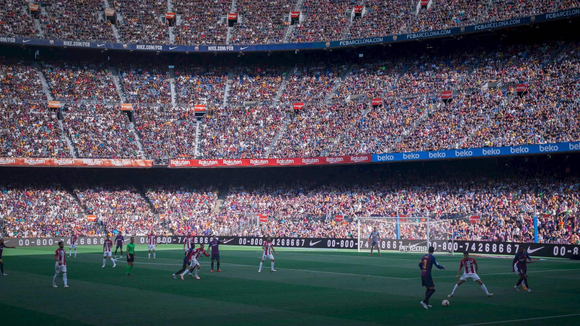 A large crowd watches a soccer match in a stadium, with players in action on the field and vibrant seating areas filled with spectators.