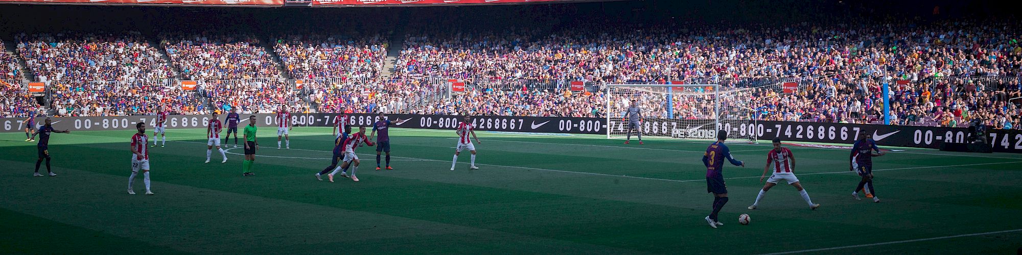 A large crowd watches a soccer match in a stadium, with players in action on the field and vibrant seating areas filled with spectators.