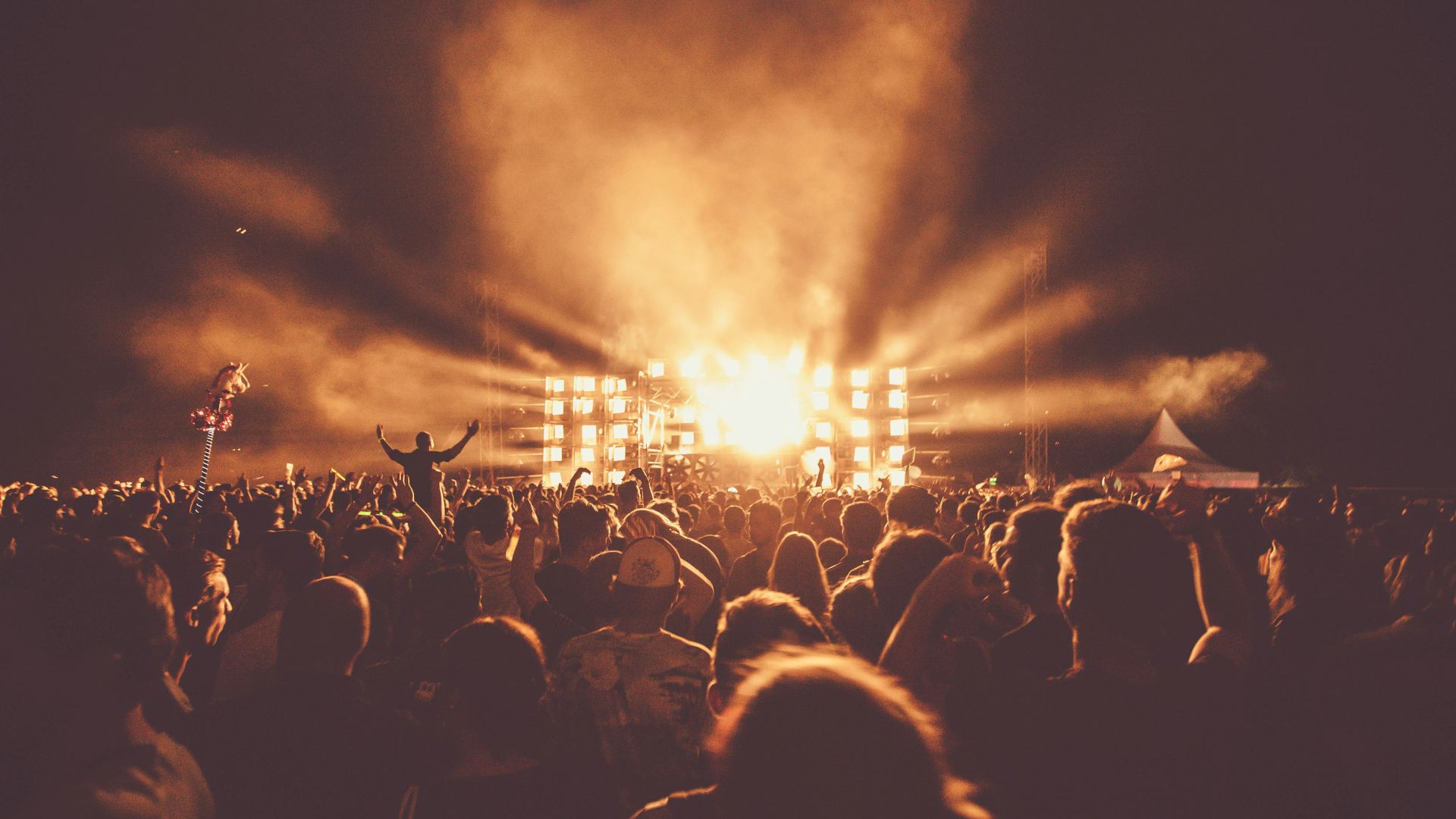 A large crowd enjoys an outdoor concert at night, with bright stage lights and haze, creating an energetic and vibrant atmosphere.