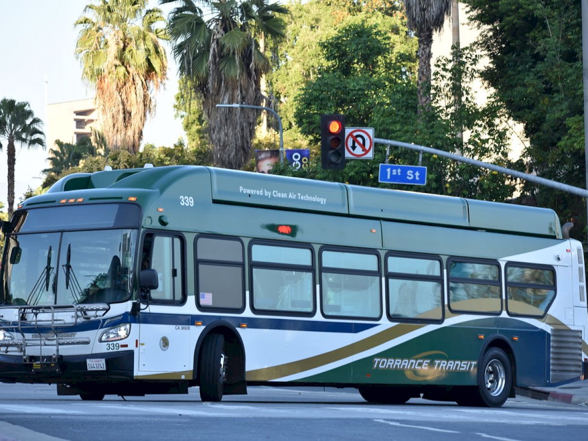 A green and white Torrance Transit bus is turning at the intersection of 1st St. and Medford, with palm trees and traffic lights in the background.