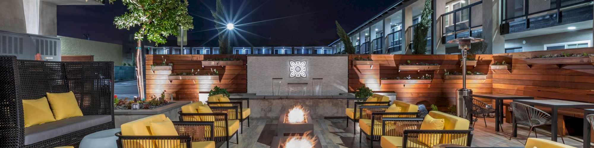 An outdoor patio at night with yellow-cushioned chairs around rectangular fire pits, surrounded by modern buildings and a lit tree.