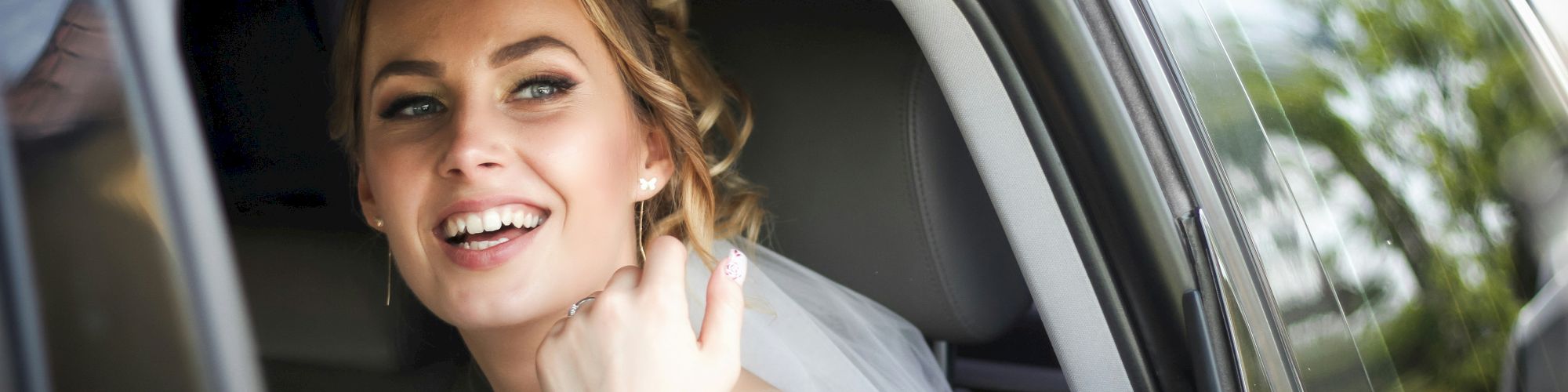 A bride smiling through an open car window, wearing a white dress with lace details. She appears joyful and the scene is bright with greenery.