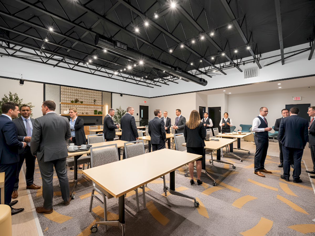 A group of people in business attire are networking in a conference room with tables and chairs, under bright ceiling lights.