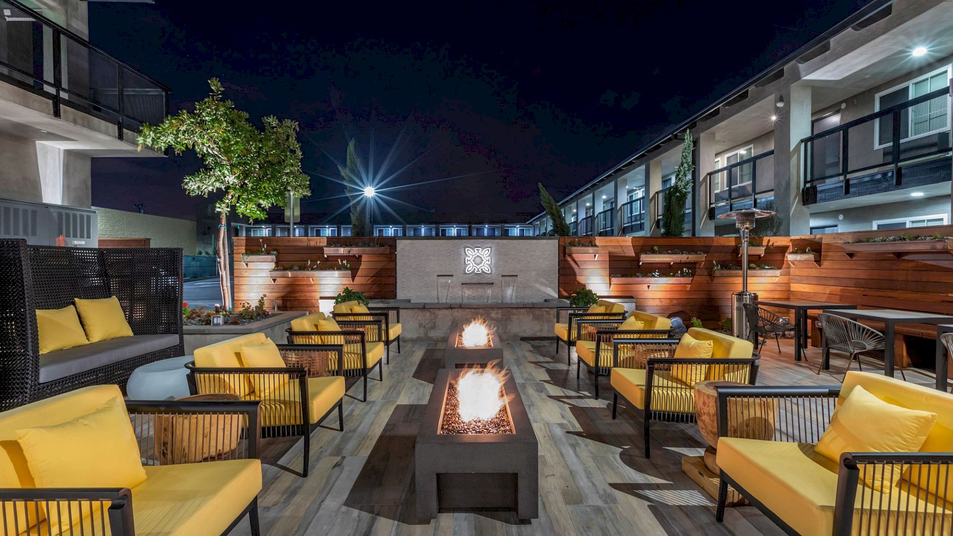 Nighttime patio courtyard with modern buildings, yellow cushioned chairs, and blazing fire pits under spotlights.