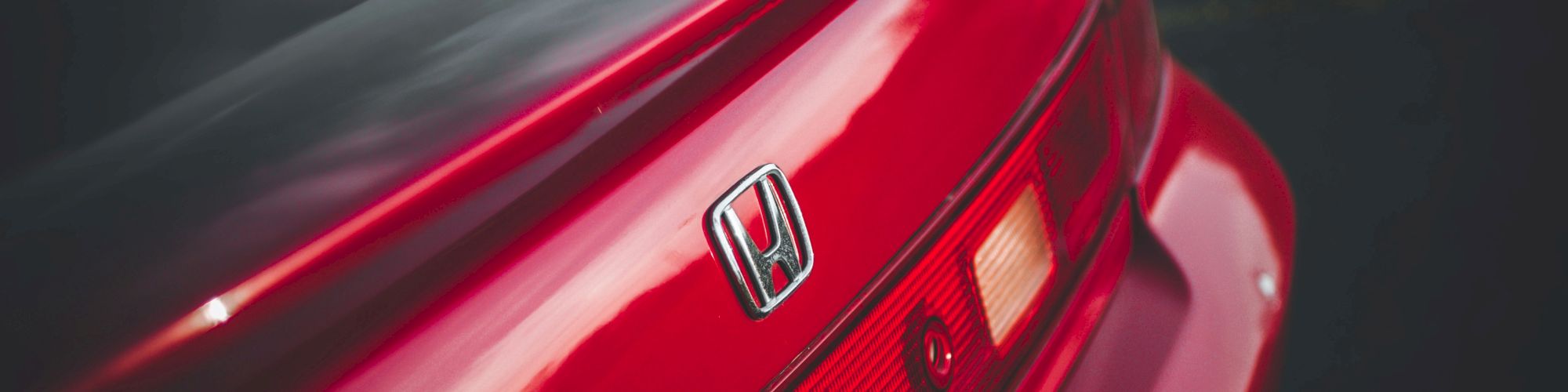 A close-up of a shiny red car’s rear, showing the Honda emblem on the trunk and its taillights, with reflections on the glossy surface.