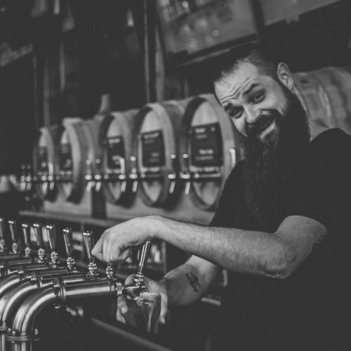 A man with a beard is smiling while pouring a drink from one of several taps, with barrels in the background, in a black and white photo.