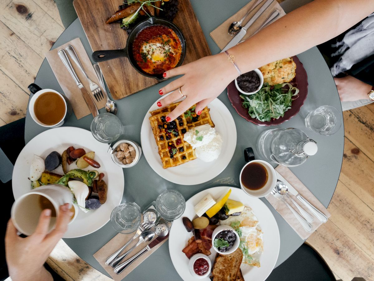 A top-down view of a table with various meals, including waffles, shakshuka, and salads, being shared among four people in a casual dining setting.