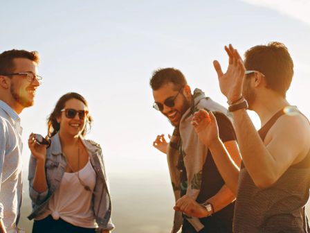 Four people are outdoors enjoying a sunny day, talking and laughing while wearing casual clothes and sunglasses.