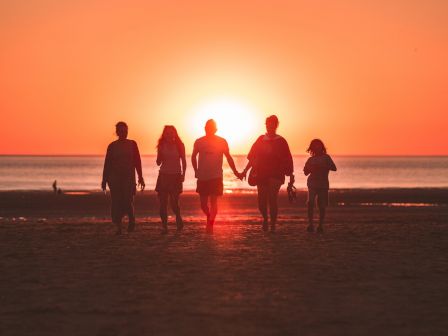 A group of five people walking on a beach at sunset, holding hands, with the sun casting a warm glow behind them, creating a serene silhouette.